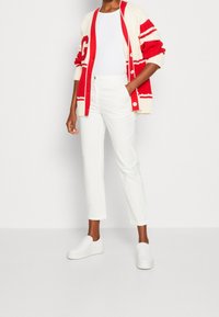 Röd och creamfärgad stickad cardigan med stor bokstav, vit t-shirt, vita skräddarsydda byxor och vita slip-on sneakers. Släta texturer och rena linjer.