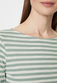 Eine Frau trägt ein grün-weißer, horizontal gestreifter Pullover, der ihren Hals, ihre Lippen und einen Teil ihrer braunen Haare zeigt.