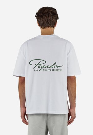 Muškarac u bijeloj majici s tekstom "Pegador All Rights Reserved" zelenom bojom na leđima, svijetlosivim hlačama, stoji okrenut leđima.