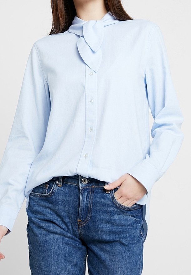 Personne portant une chemise bleue claire boutonnée avec un détail de cravate au col et un jean bleu avec une main dans la poche.