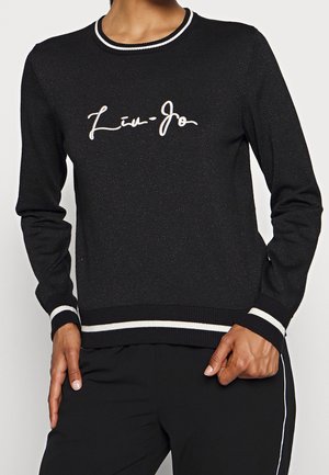 Woman wearing a black sweater with white trim and cursive "Liu Jo" logo, paired with black pants featuring white side stripes.