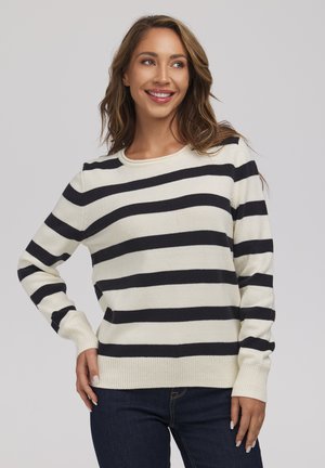 Kobieta uśmiechająca się, ubrana w biały sweter z szerokimi czarnymi poziomymi paskami i ciemnoniebieskie dżinsy, pozująca na tle gładkiego jasnego tła.