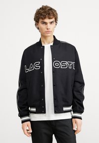 Svart bomberjacka med vitt kontrasterande logo "LACOSTE." Har randiga muddar och halsringning. Tillverkad av ett smidigt tyg.