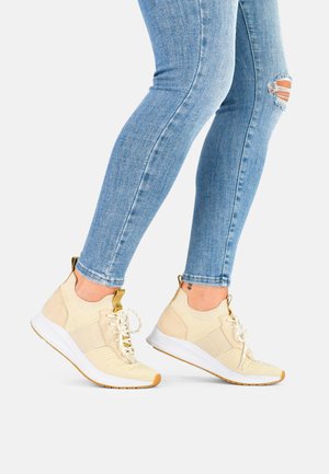 Beige high-top sneakers with textured fabric and knotted laces, featuring a white rubber sole and a yellow accent on the toe, paired with blue jeans.