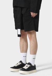 Black cargo shorts with front and side pockets, featuring a stitched label on the waistband. Worn with white socks and black sneakers.