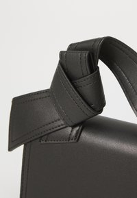 Bolso de mano de cuero negro con forma estructurada, acento de asa plegada y costuras visibles. Textura suave con un acabado mate.
