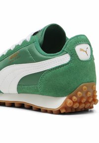 Zapato deportivo verde y blanco con parte superior de ante y nylon, suela de goma con púas circulares y acento de logo dorado en el lateral.