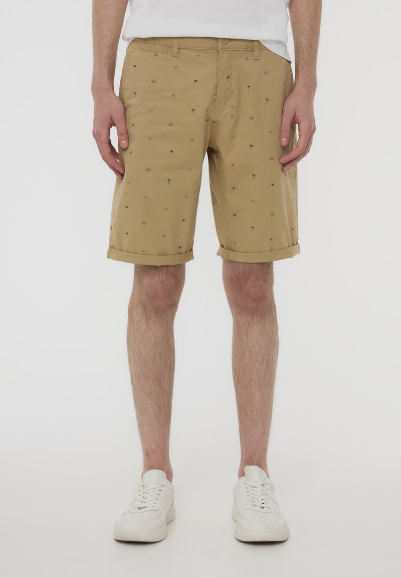 Pantaloni beige con risvolti, caratterizzati da un motivo di piccole palme e forme geometriche, realizzati in tessuto leggero, con tasche frontali visibili.