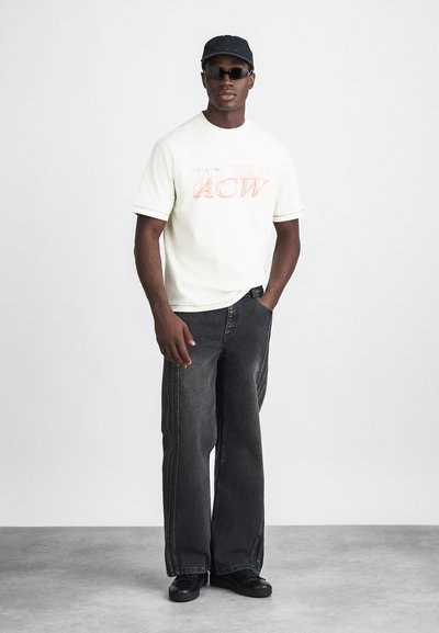 Camiseta blanca con texto rojo "ACW", combinada con jeans negros de pierna ancha. Una gorra negra y gafas de sol completan el look. Tela suave, diseño casual.
