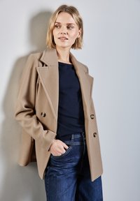 Beige villakangastakki, jossa on kaksirivinen muotoilu, suuret lovilaput ja kultaiset napit, yhdistettynä tummansiniseen neuleeseen ja tummansinisiin farkkuihin.