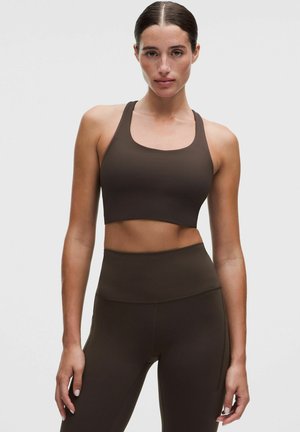 Ung kvinde med bag slicket hår iført en mørkebrun sports-bh og høj-taljede leggings, der står mod en ensartet lys baggrund.