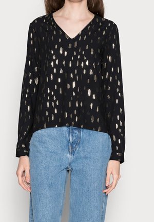 Kobieta ubrana w czarną bluzkę z długim rękawem i dekoltem w serek z pionowymi metalicznymi złotymi paskami oraz jasnoniebieskie jeansy z denimu.