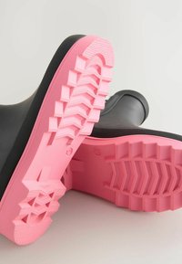 Des bottes en caoutchouc noires avec une surface texturée présentent des semelles roses vif, à motifs et une prise rigide pour une meilleure traction.