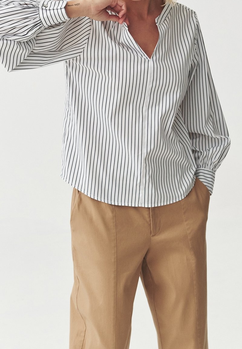 Henkilö, joka pukeutuu valkoiseen pitkähihaiseen paitaan, jossa on ohutta mustaa pystysuoraa raitaa, ja beigeihin housuihin. Yksi käsi on taskussa ja toinen lähellä kasvoja.