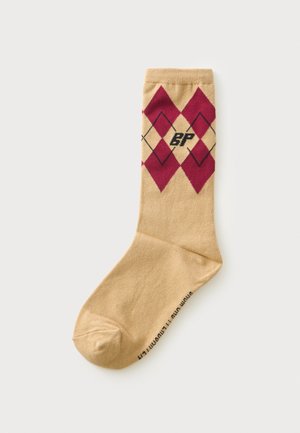 Beige sok met rood en zwart ruitpatroon en zwarte tekst nabij de teen op een effen lichte achtergrond.