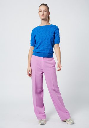Blauer kurzärmeliger Strickpullover mit Wellenmuster, kombiniert mit hochtaillierten rosa Hosen und metallischen Sneakers, vor einem neutralen Hintergrund stehend.
