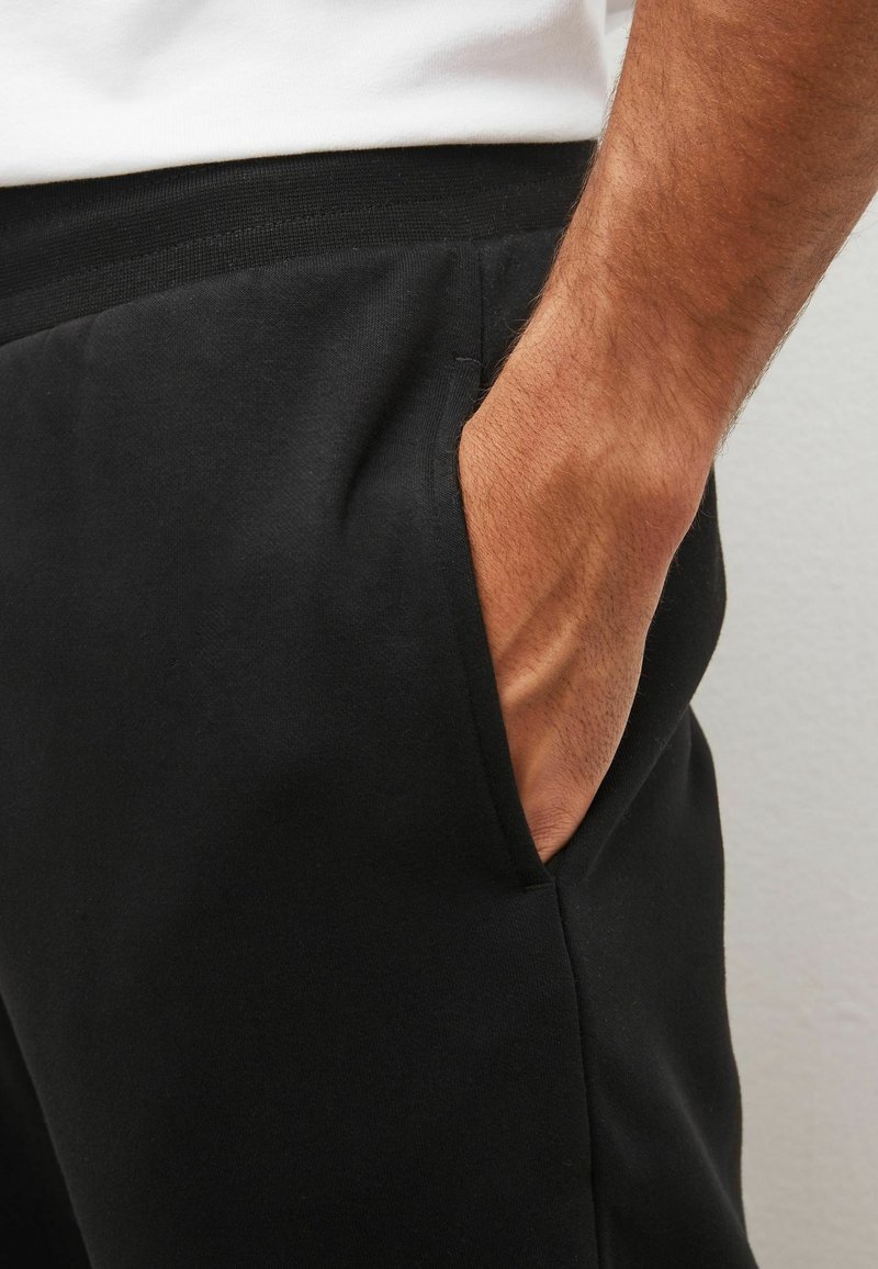 Black sweatpants featuring an elastic waistband, side pockets, and a soft cotton blend texture, shown from the waist to the pocket.