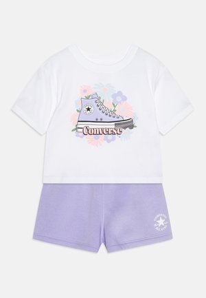 Valkoinen lyhythihainen t-paita, jossa on painettu laventelin värinen Converse-kengä ja kukkakuviota, yhdistettynä vaaleanlila lyhythihaisiin shortseihin, joissa on logo.