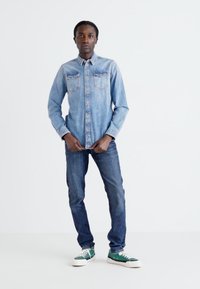 Camisa de denim en azul claro con bolsillos en el pecho, combinada con jeans azul oscuro y zapatillas verdes. De pie contra un fondo blanco.