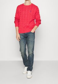 Pull rouge en maille torsadée avec poignets côtelés, porté avec un jean bleu et des baskets blanches. Comprend un petit logo sur la poitrine.