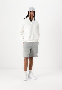 Felpa bianca con zip, pantaloni corti grigi con il logo Nike bianco, cappellino grigio e sneaker bianche. Abbigliamento sportivo casual con una vestibilità rilassata.