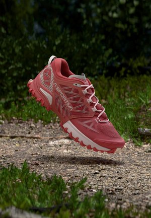 Chaussure de trail rouge avec semelle texturée flottant au-dessus d'un chemin de gravier entouré de végétation verte.