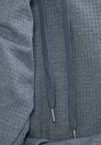 Harmaa teksturoitu huppari, jossa on kudottu kuvio, tummat nyörit metallipäillä ja pehmeä kangaspinta.