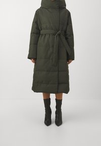Dlouhý zeleno-olivový péřový kabát s páskem, kapucí a širokými rukávy. Texturovaná látka má vodorovné prošívání a měkký povrch.