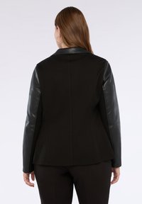 Vrouw met lang bruin haar draagt een zwarte jas met leren mouwen, weergegeven van achteren tegen een witte achtergrond.