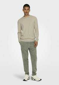 Beige langemouwen trui met geribbelde boorden, gecombineerd met groene corduroy broek. Model draagt een crème sneakers met donkere accenten, staand voor een neutrale achtergrond.