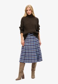 Pull à col roulé en tricot marron, jupe évasée à carreaux bleus avec des accents marron et or, complétée par des bottes à talons beiges.