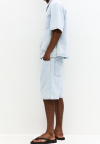 Světlomodrá texturovaná košile s krátkým rukávem a odpovídající šortky, obě s kapsami, doplněné černými žabkami.