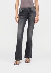 Γυναίκα που φοράει ξεθωριασμένο μαύρο τζιν με καμπάνες και σανδάλια με τακούνι και λουριά, στέκεται μπροστά σε απλό λευκό φόντο.