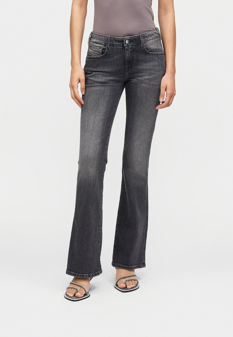 Γυναίκα που φοράει ξεθωριασμένο μαύρο τζιν με καμπάνες και σανδάλια με τακούνι και λουριά, στέκεται μπροστά σε απλό λευκό φόντο.