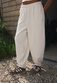 Smėlio spalvos platūs sportinės kelnės su elastinga juosmens juosta, šoniniais kišenėmis ir suvarstytais kulkšniais, derinamos su tamsiai ruda sportbačiais su baltomis juostelėmis.