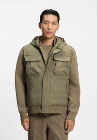Oliven grøn hættevest med to frontlommer, lynlås lukning og ribbet kant. Bæres over en beige top med en glat, mat struktur.