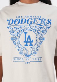 Lys grå T-shirt med blåt grafisk tekst "LOS ANGELES DODGERS," en baseball og dekorative mønstre; rund hals, korte ærmer.