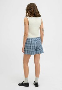 Witte gebreide mouwloze top en blauw-witte geruite shorts. Persoon met de rug naar de camera, draagt grijze sokken en zwarte schoenen.