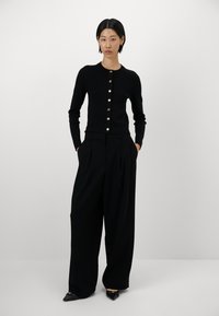 Sorte cardigan med rund hals og guldknapper, parret med brede sorte bukser og spidse sorte sko, på en neutral baggrund.