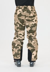 Camouflage skibroek in tinten van beige en groen, met een ruitpatroon, waterdicht materiaal en gearticuleerde knieën voor beweging.
