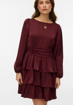 Vīna sarkanas krāsas kleita ar garām piedurknēm, savilktu vidukli un daudzslāņu kroku svārku. Gluda faktūra un smalka spīduma efektu. Valkāts zelta kulona kaklarota.