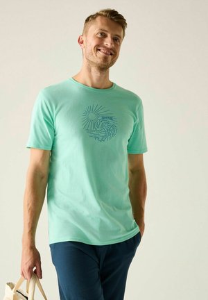 Smiling man wearing a light green T-shirt with a sun and wave design, walking with one hand in pocket and holding a beige bag in the other hand.