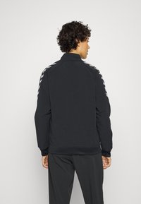 Svart zip-up jacka med ståkrage, med kontrasterande vita randiga detaljer på axlarna samt ribbad nederdel och ärmslut.