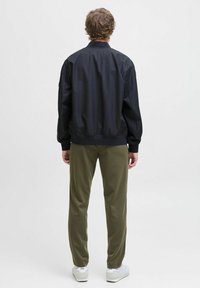 Veste aviateur noire avec une texture lisse, des poignets côtelés et un col montant, associée à un pantalon vert olive et des baskets blanches.