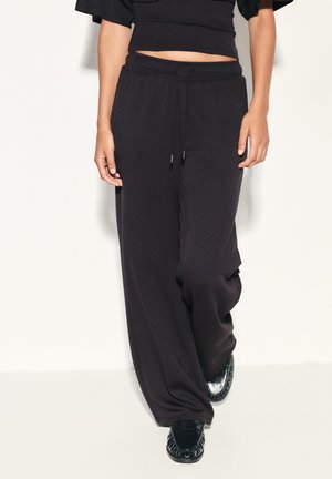 Personne portant un pantalon de survêtement noir ample avec cordon et des chaussures noires brillantes style mocassin, marchant sur un fond clair.