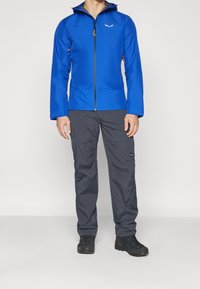 Blå vattentät jacka med dragkedja och hög krage, kombinerad med mörkgrå cargobyxor med fickor och förstärkt sömnad. Svarta skor.