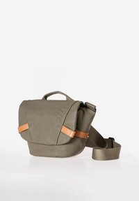 Borsa a tracolla compatta di colore verde oliva con tracolla regolabile, manico superiore, chiusura a patta e cinghie di accentatura in pelle beige su sfondo bianco.