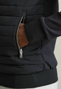 Geacă neagră căptușită cu un buzunar lateral cu fermoar. Include manșete și tiv ribbed, textură de țesătură netedă și detalii cu fermoar metalic.