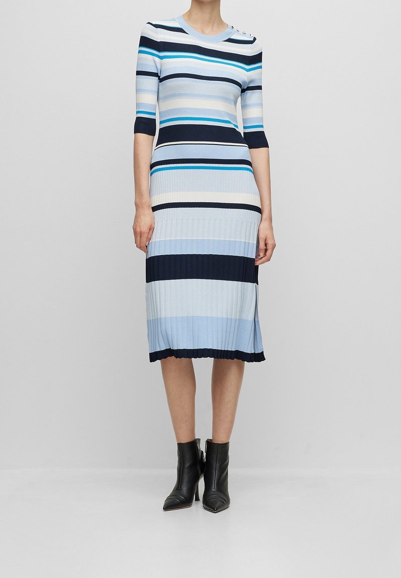 Femme portant une robe côtelée longueur genou à rayures horizontales bleu et blanc, avec des bottines noires, debout devant un fond uni.