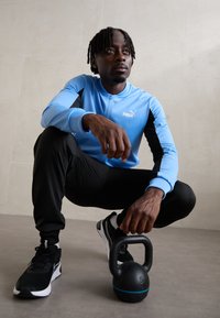 Costumul sportiv Puma prezintă o jachetă bleu deschis cu fermoar și accente negre, pantaloni negri și adidași negri, cu o greutate kettlebell pe podea.